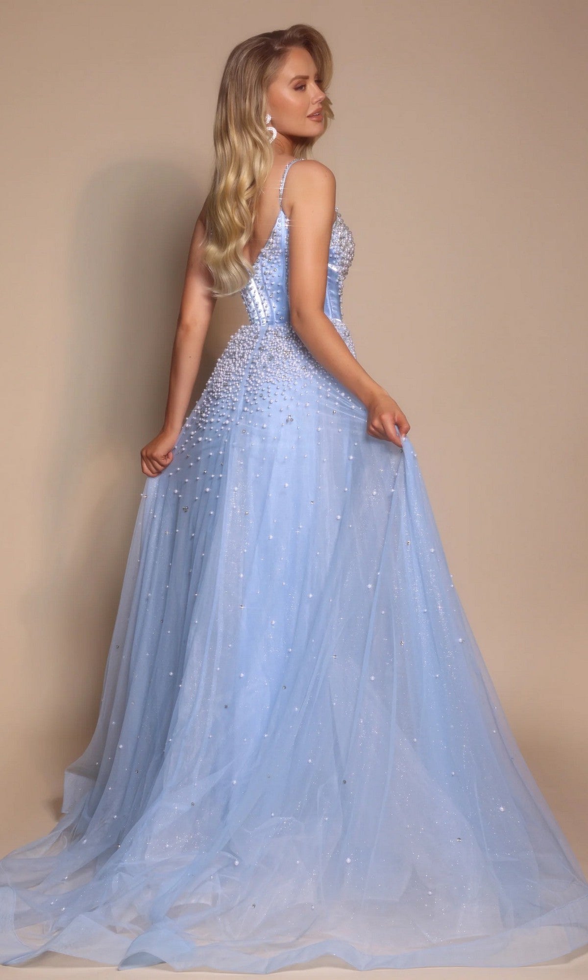 A woman with long blonde hair poses against a beige background wearing the Corseted Ball Gown: Portia and Scarlett PS26208, a light blue, floor-length gown with a sparkly boned-corset bodice, thin straps, and pearl embellishments.